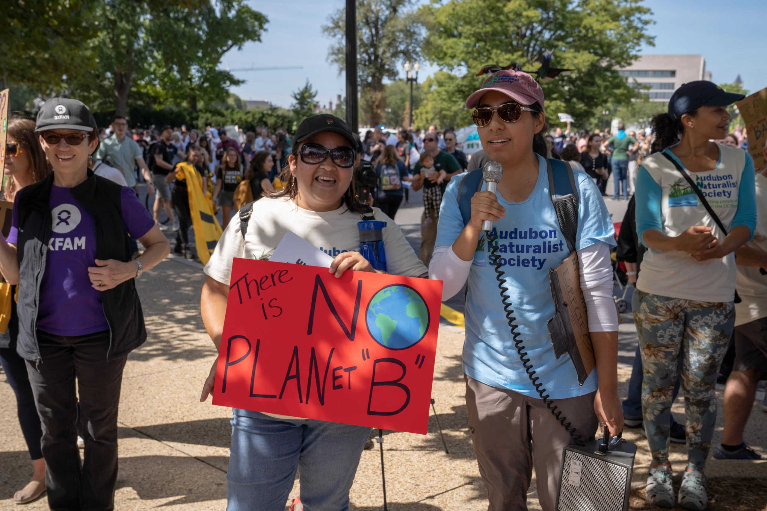 Two Latina women stand in the midst of a crowd at a march on an autumn day in Washington, DC. On the left, one holds a sign saying "There is No Planet B". On the right, the other holds a megaphone to rally the crowd.