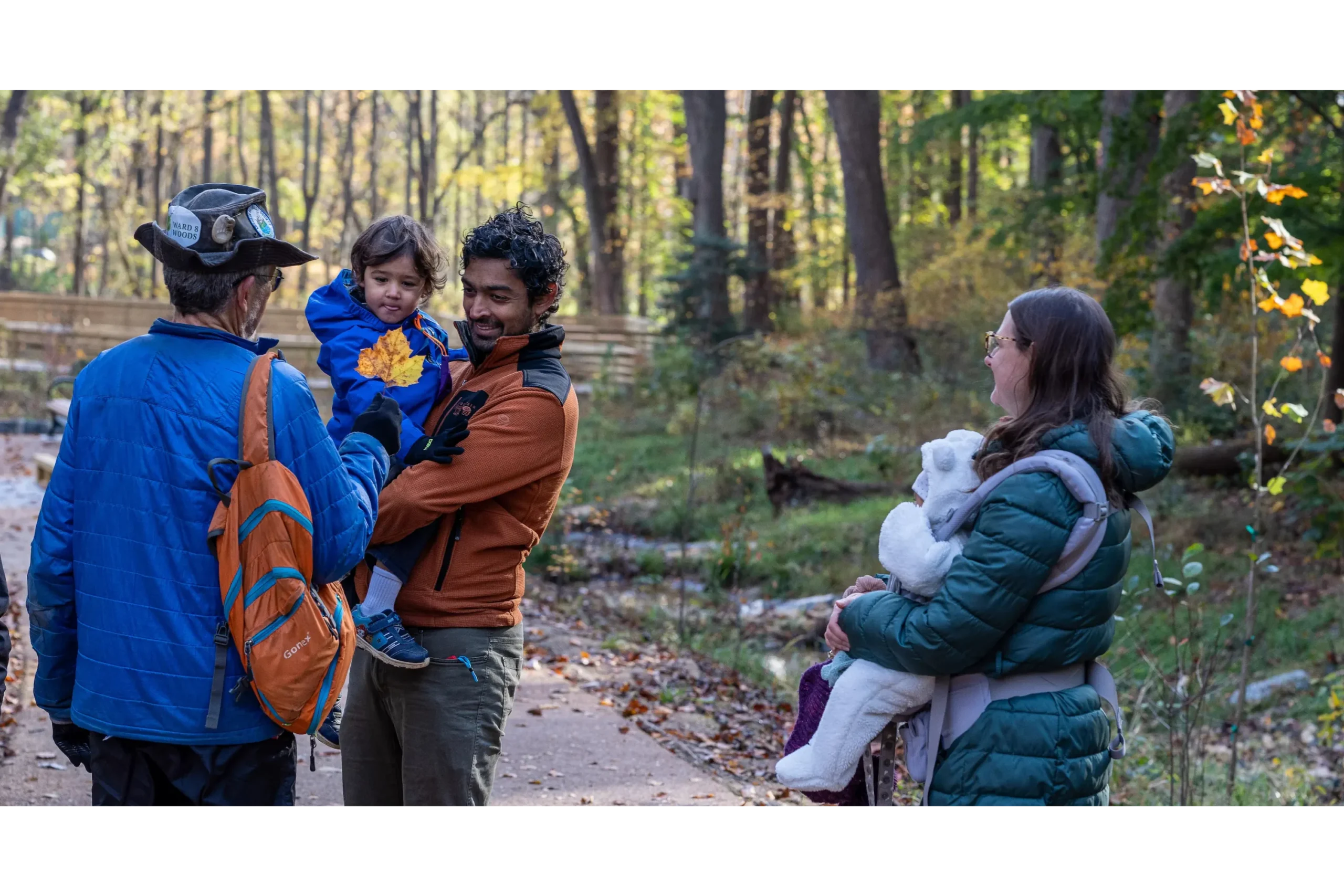 A guide shows a large fall leaf to a toddler in his smiling father's arms while a mother and baby in sling look on