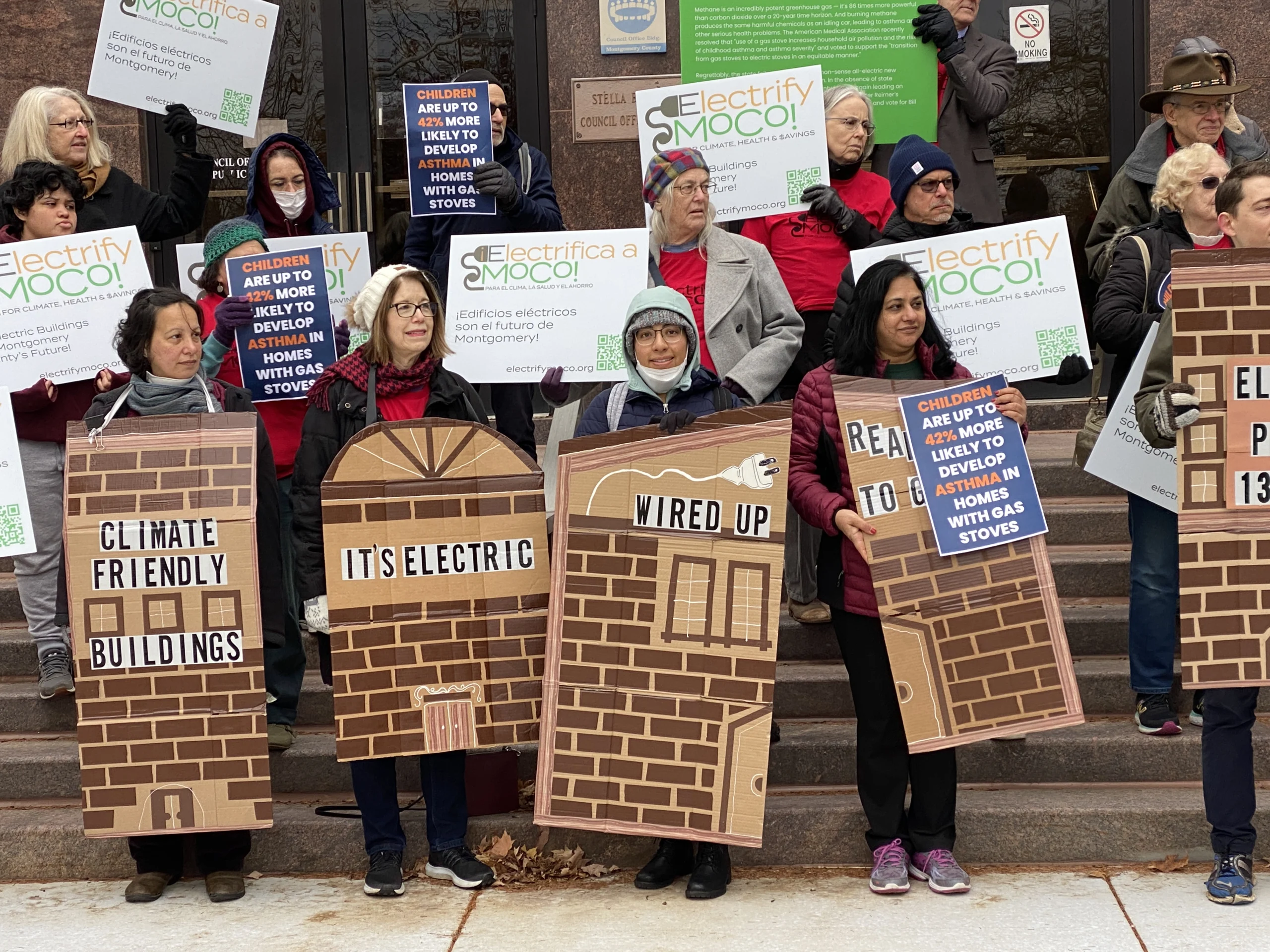 Four people holding cardboard signs made to look like buildings stand in the front row of a rally on the steps of the Montgomery County Council building. It is a chilly day in November and people are wearing hats and coats. On the buildings they say "Climate Friendly Buildings" "It's Electric" "Wired Up" "Ready to Go".