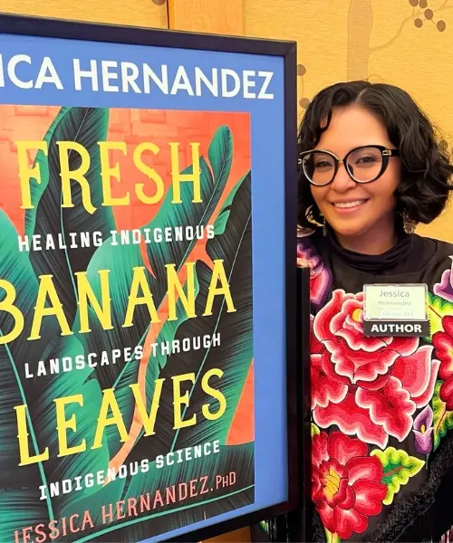 Dr. Jessica Hernandez poses alongside a poster images of her book Fresh Banana Leaves: Healing Indigenous Landscapes through Indigenous Science.