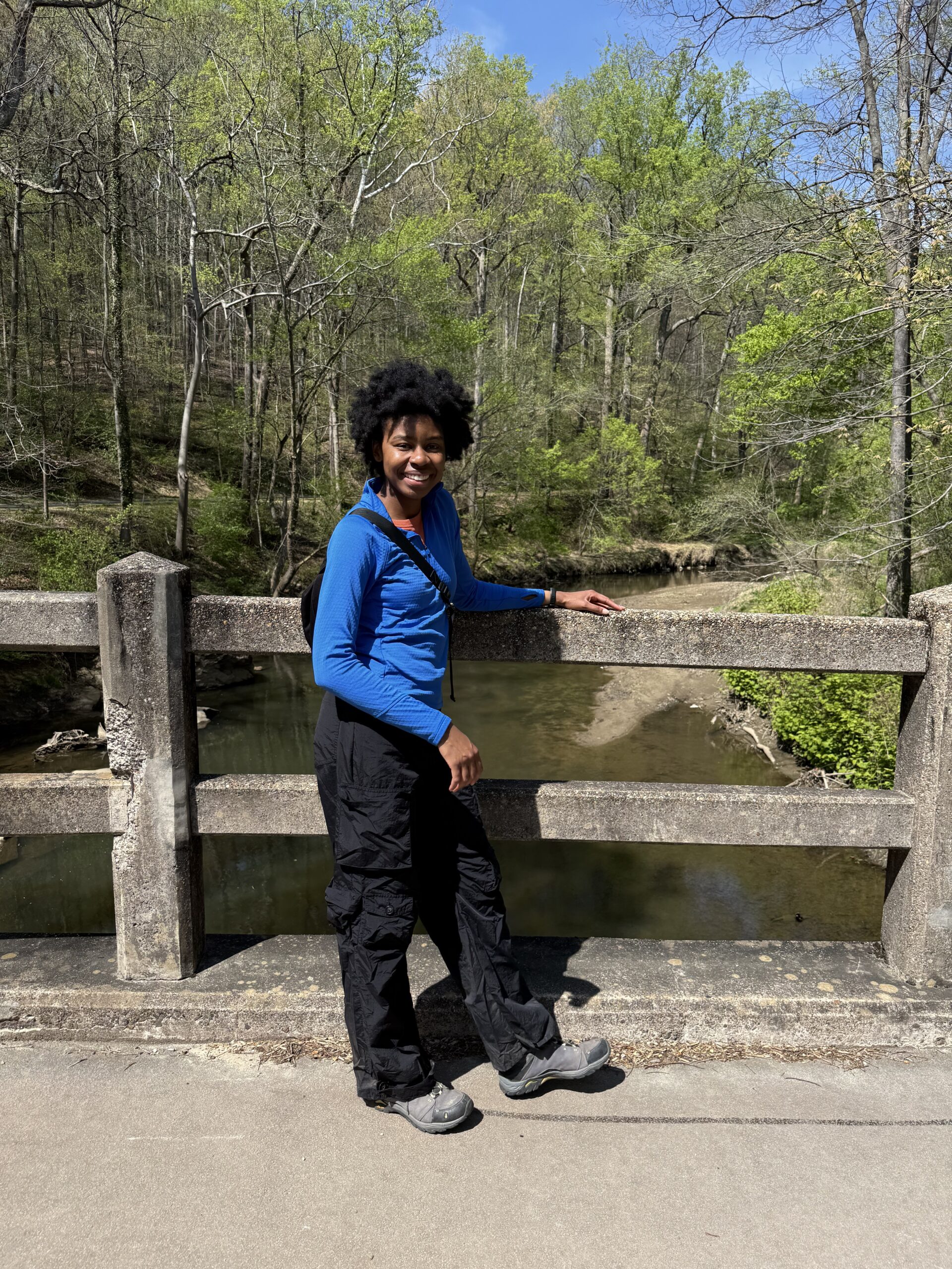 Black woman standing up right in a blue quarter zip, black hiking pants, and hiking boots, posed on a bridge in Rock Creek Park with a stream flowing behind.