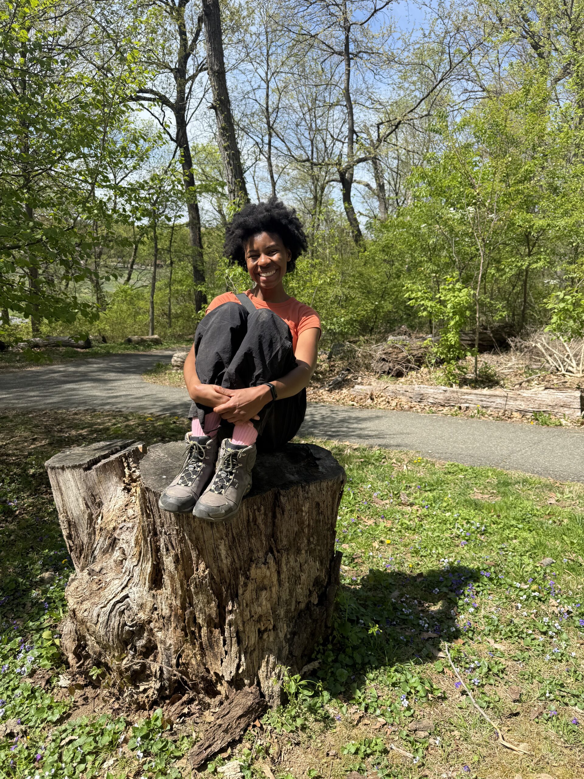 Black woman in an orange short sleeve top with with black pants sitting on a tree stump, smiling.