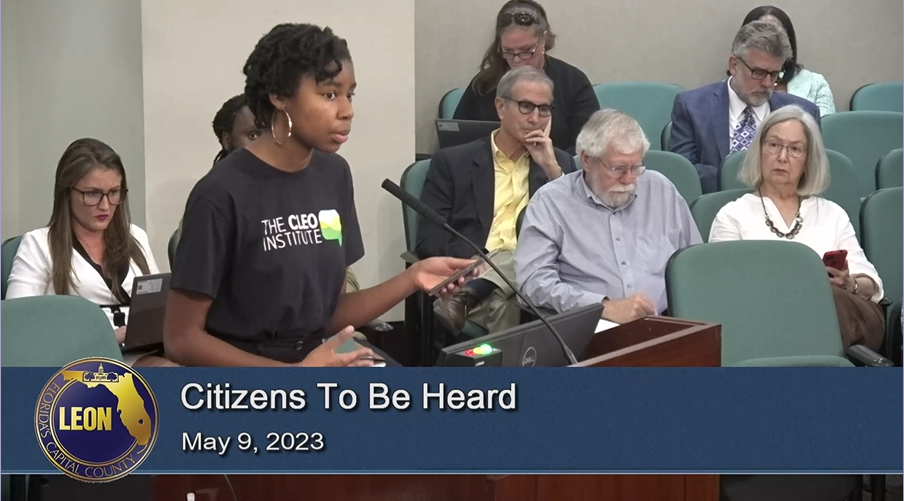 Black woman with a short twisted hair style and large hoop earrings standing in front of a microphone in a county commission office. She is wearing a shirt that says "The CLEO Institute." She is giving public testimony before the Leon County Commission meeting on May 9, 2023 during public comment period entitled "Citizens To Be Heard."